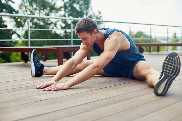Young sporty man with closed eyes doing stretching exercise and enjoying fresh air while sitting on wooden floor of spacious terrace, picturesque view on background - Photo, Image