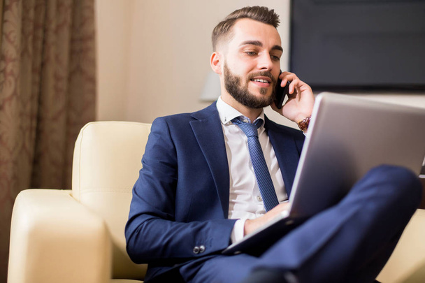 Portrait of handsome young entrepreneur speaking by phone and using laptop while working in comfortable hotel room or office - Foto, afbeelding