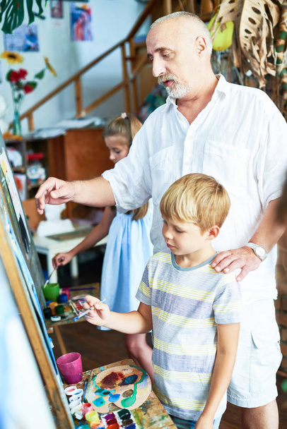 Portrait of senior teacher helping little boy painting picture in art studio standing by easel with other children in background - Photo, Image