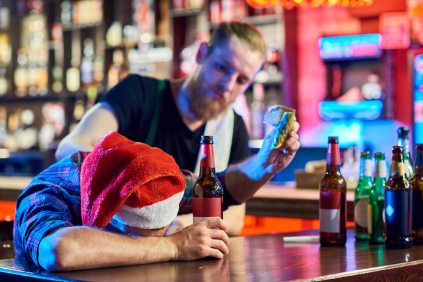 Portrait of drunk man wearing Santa hat laying on bar counter drinking beer and throwing money around after Christmas party in pub - Photo, Image