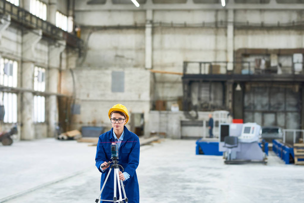 Portrait of confident worker wearing uniform and hardhat making measurements with help of total station theodolite while standing at spacious industrial workshop - Photo, Image