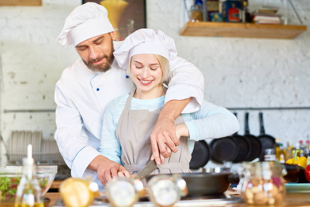 Confident bearded chef wearing uniform helping his pretty young assistant to cut vegetables while working together at modern restaurant kitchen - Фото, зображення