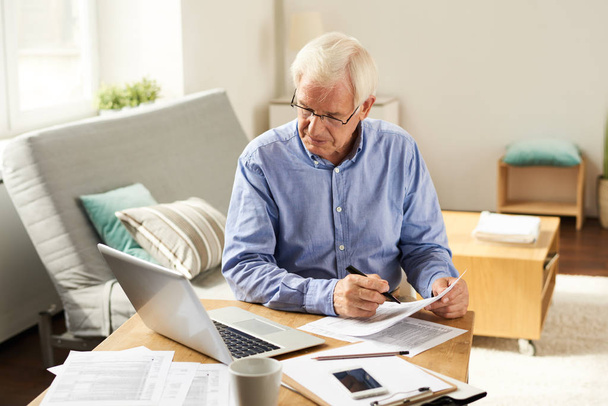 Retrato del hombre mayor moderno llenando el formulario de solicitud en casa sentado en la mesa con el ordenador portátil en la sala de estar
 - Foto, imagen