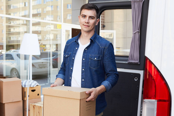 Portrait of young man holding cardboard box and smiling happily to camera  while unloading moving van outdoors - Photo, Image
