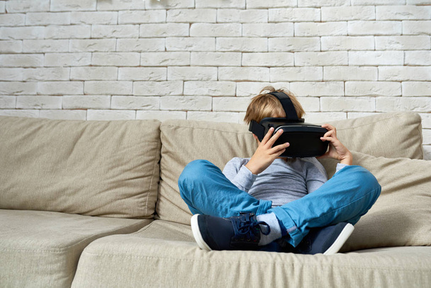 Täyspitkä muotokuva pikkupoika yllään VR lasit istuu rajat jalat sohvalla, kopioi tilaa
 - Valokuva, kuva