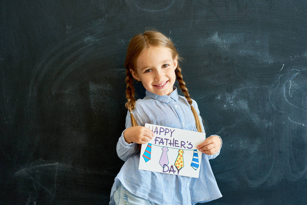 Portrait of smiling little girl holding handmade  greeting card for Fathers Day posing against blackboard in school and looking at camera - Foto, Bild