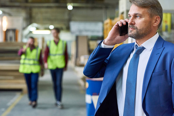 Portrait of handsome mature businessman speaking by phone in factory workshop with two workers in background, copy space - Foto, Bild