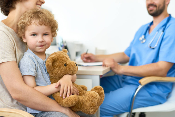 Portrait of cute child sitting on mothers lap in doctors office hugging plush teddy bear  and smiling at camera, copy space - Photo, Image