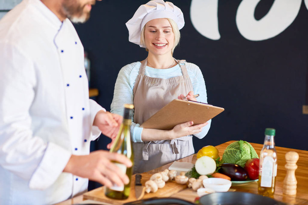 Πορτρέτο της χαμογελαστός νεαρός βοηθός φορώντας ποδιά είναι απαραίτητο σημειώσεις ενώ γενειοφόρος σεφ δείχνει πώς να προετοιμάσει σωστά το πιάτο, εσωτερικό του σύγχρονο εστιατόριο κουζίνα σε φόντο - Φωτογραφία, εικόνα