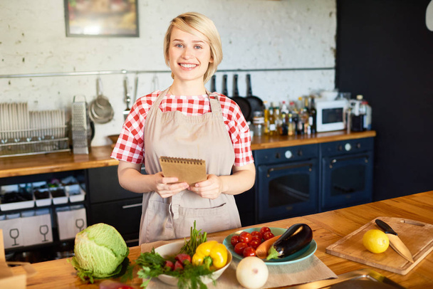 Cheerful blond-haired housewife wearing checked shirt and apron looking at camera with toothy smile while standing at kitchen table and holding notepad with recipe in hands, waist-up portrait - Photo, Image