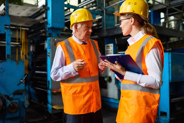 Retrato de gerente de fábrica sênior conversando com o jovem assistente escrevendo instruções, ambos vestindo chapéus duros e jaquetas reflexivas
 - Foto, Imagem