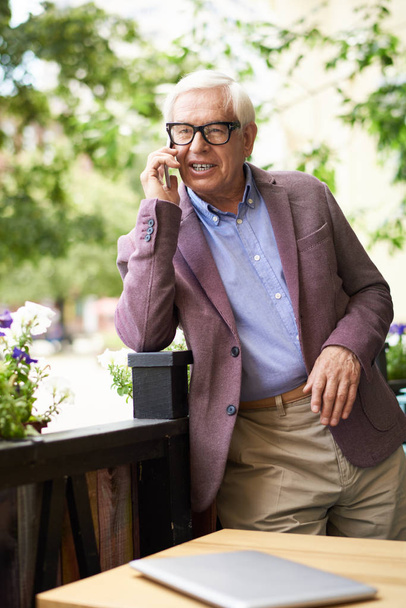Portret van moderne senior man spreken via de telefoon buitenshuis in café, leunend op flowerbed - Foto, afbeelding