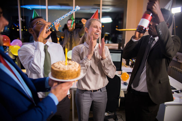 Young businesspeople celebrating birthday of coworker in office, blowing party horns, drinking and having fun - Photo, Image