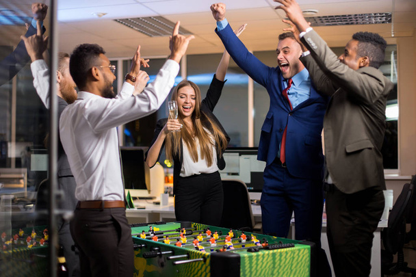 Νέοι επιχειρηματίες χαρούμενα στέκεται με επιτραπέζιο ποδοσφαιράκι και πανηγυρισμούς νίκη ενώ Εταιρικό πάρτι στο γραφείο - Φωτογραφία, εικόνα