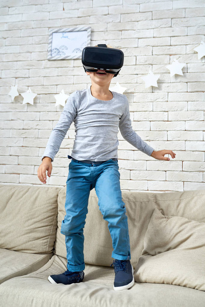 Портрет збудженого маленького хлопчика, який стоїть на дивані вдома в окулярах VR, щоб дивитися 360 відео і грати в ігри доповненої реальності
 - Фото, зображення