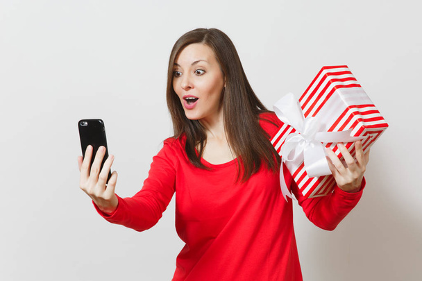 Γυναίκα κάνει selfie στο κινητό τηλέφωνο με κόκκινο ριγέ παρόν πλαίσιο με δώρο που απομονώνονται σε λευκό φόντο. Αντιγράψτε το χώρο για τη διαφήμιση. Ημέρα του Αγίου Βαλεντίνου, ημέρα της γυναίκας, έννοια Ενοικιαζόμενα. - Φωτογραφία, εικόνα