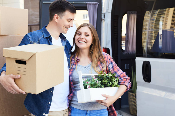 Portrait of happy  young couple embracing and smiling at camera holding cardboard boxes next to moving van outdoors - Photo, Image