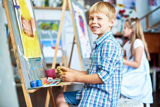 Side view portré kis fiú zsírkréták ház festőállvány a művészeti stúdió és látszó-on fényképezőgép, boldogan mosolygó - Fotó, kép