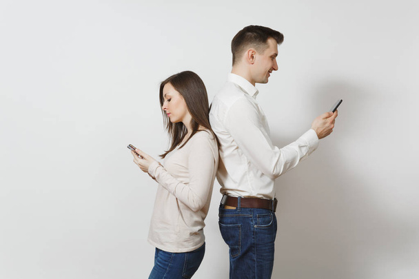 Young caucasian couple. Man and woman in love in casual light clothes, jeans standing back to back, using and texting on their mobile smart phones isolated on white background. Gadget today concept. - Foto, Imagen