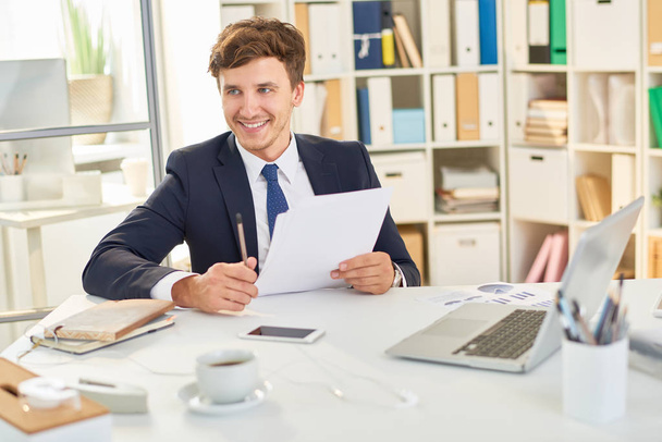 Portrait of smiling young businessman working at desk in office, reading documentation - Fotó, kép