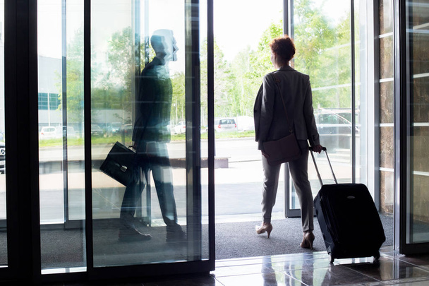 Back view silhouette of young businesswoman with suitcase walking through sliding glass doors of hotel, copy space - 写真・画像