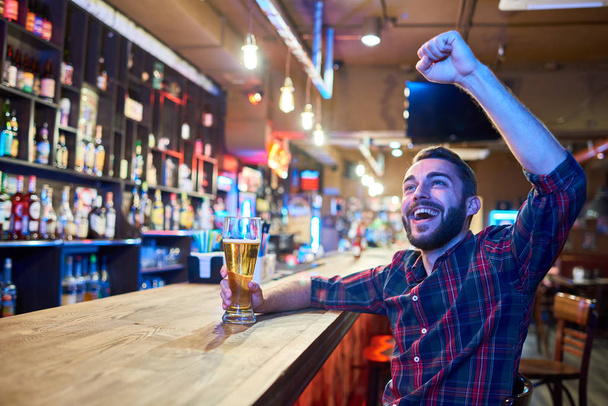 Portrait of excited sports fan cheering happily in pub watching match on TV raising hands and drinking beer, copy space - Фото, изображение