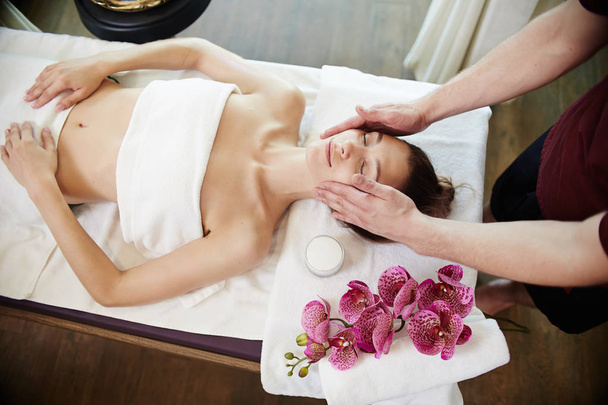 High angle portrait of young woman enjoying face lifting massage lying on table in SPA center and relaxing, smiling blissfully - Photo, Image