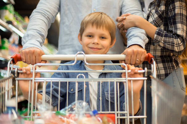 Πορτρέτο του happy νεαρό αγόρι αγορές για ψώνια στο σούπερ μάρκετ μαζί με τους γονείς, πιέζει το καλάθι και χαμογελά στη φωτογραφική μηχανή - Φωτογραφία, εικόνα