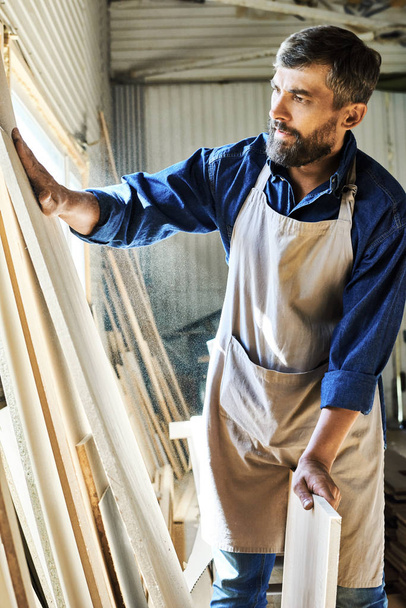 Porträt eines gelernten Tischlers, der im Atelier ein glattes Stück Holz sanft berührt und die besten Materialien auswählt - Foto, Bild