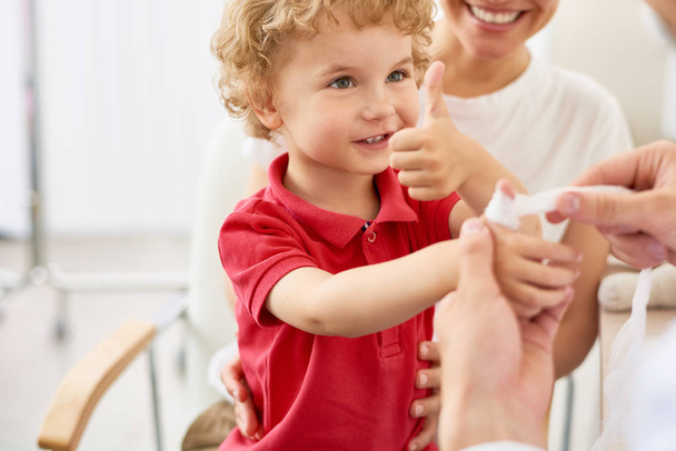 Portrait of adorable curly child showing thumbs up at doctors office during visit with mom - Foto, Imagem