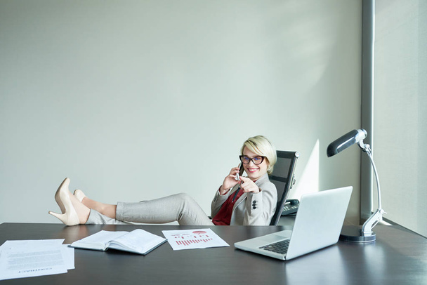 Full length portrait of cheerful young businesswoman sitting with feet on office desk while talking to her friend on smartphone, she pointing at something with index finger - Photo, Image