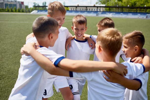 Ομάδα από αγόρια με τη στολή αγκαλιάζει χαμογελώντας ευτυχώς κατά τη διάρκεια της πρακτικής στο γήπεδο ποδοσφαίρου - Φωτογραφία, εικόνα