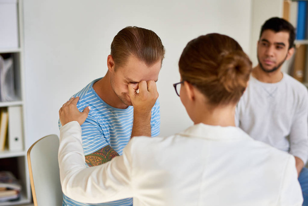 Portret van moderne jonge man huilen terwijl het delen van problemen met vrouwelijke psychiater in groepstherapie sessie - Foto, afbeelding