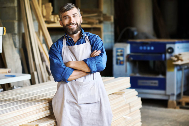 Portret van volwassen bebaarde timmerman poseren met vertrouwen met armen gekruist permanent in houtbewerking winkel en lacht vrolijk kijken camera - Foto, afbeelding