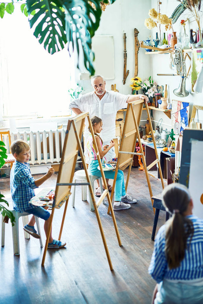 Ryhmä lahjakkaita lapsia istuu edessä easels ja luoda asetelma maalauksia, niiden vanhempi opettaja pitää silmällä niitä, sisätilojen tilava taidestudio taustalla
 - Valokuva, kuva