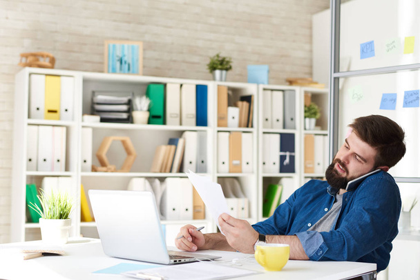 Ritratto di uomo barbuto moderno in abbigliamento casual mentre parla per telefono al lavoro in ufficio moderno
 - Foto, immagini