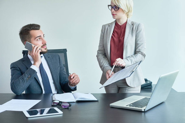 attraktive blondhaarige Managerin, die ihrem hübschen männlichen Kollegen Finanzzahlen zeigt, während er mit ihrem Chef telefoniert, im Hintergrund Großraumbüro - Foto, Bild