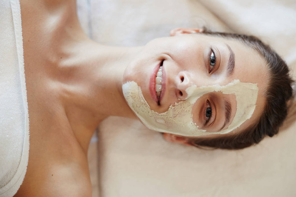 Widok z góry zbliżenie portret błogi młoda kobieta korzystających zabiegi kosmetyczne i maski na twarz w Spa, leżącego na stole do masażu na szczęście uśmiecha się do kamery - Zdjęcie, obraz