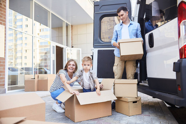 Portrait of happy young family with little son loading cardboard boxes into moving van outdoors - Photo, Image