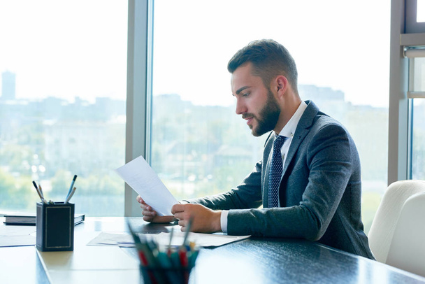 Profile view of concentrated young white collar worker wrapped up in paperwork while sitting at desk, interior of modern office with panoramic windows on background - Φωτογραφία, εικόνα