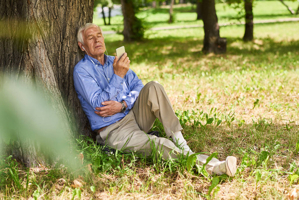 Portret van moderne senior man ontspannen luisteren naar muziek van smartphone in park onder boom - Foto, afbeelding