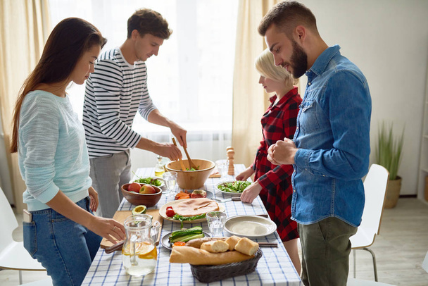 Группа молодых людей готовит ужин к праздничному торжеству стоя за большим столом вместе накрывая еду
 - Фото, изображение