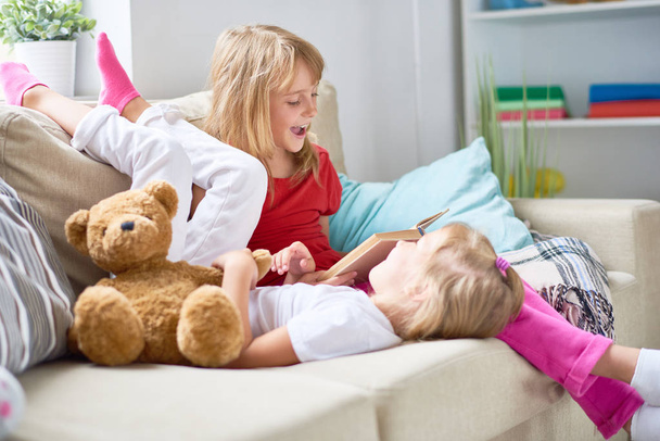 Pretty blond-haired girl reading adventure story aloud while her little sister lying on sofa and listening to her with interest, interior of cozy living room on background - 写真・画像