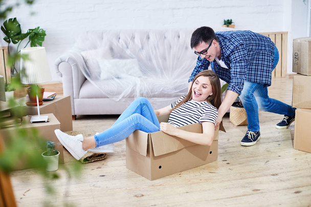 Lovely young couple having fun while riding in cardboard box at new apartment, interior design items and moving boxes on background - Φωτογραφία, εικόνα