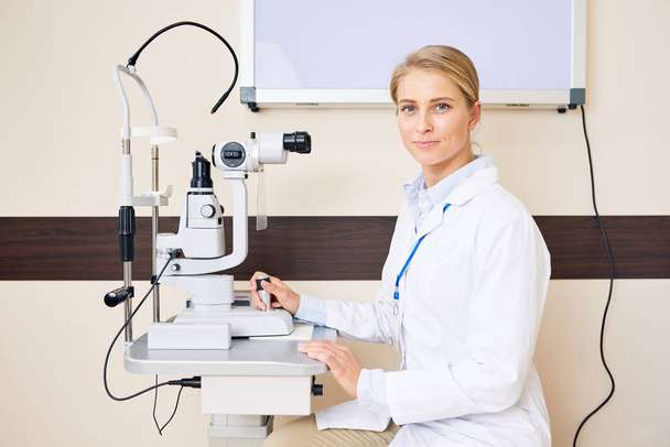 Portrait of blonde female ophthalmologist sitting at slit lamp machine looking at camera and smiling - Foto, afbeelding
