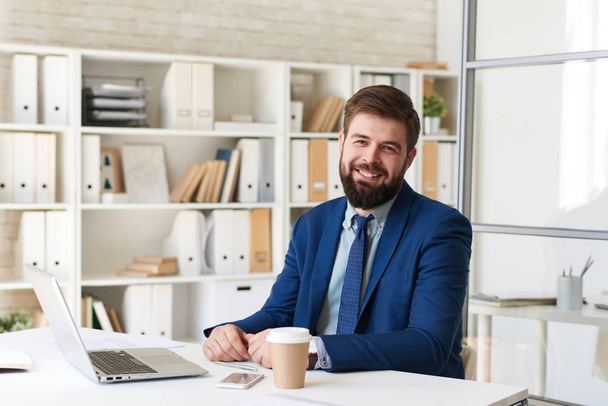 Портрет успешного бородатого бизнесмена, весело улыбающегося во время работы с ноутбуком в современном офисе
 - Фото, изображение