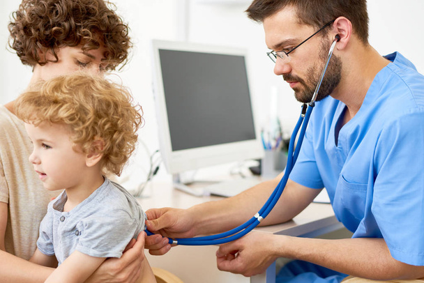 Portrét kudrnaté malého chlapce sedí na klíně matky v kanceláři lékaři zároveň pediatr, poslech dýchání pomocí stetoskopu - Fotografie, Obrázek
