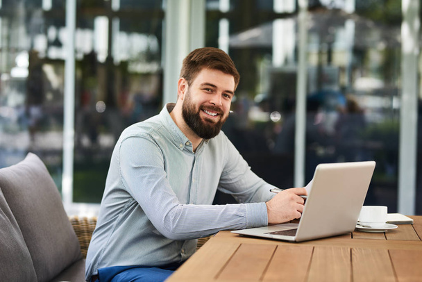 Portret udany młody biznesmen, patrząc na kamery i uśmiechając się podczas pracy z laptopem w nowoczesnym biurze - Zdjęcie, obraz