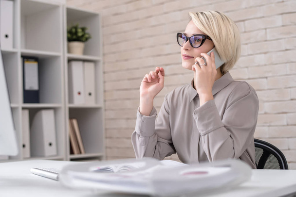 Portrait of blonde successful businesswoman speaking by phone sitting at desk in modern office and talking to client - Foto, Imagem