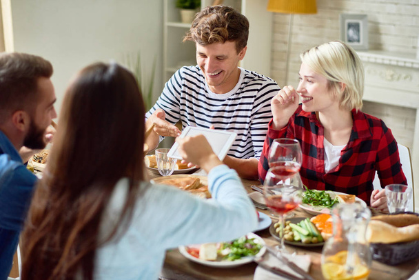 Портрет молодих людей, які сміються, щасливо дивлячись на цифровий планшет, насолоджуючись вечерею з друзями, сидячи за великим столом з їжею
 - Фото, зображення